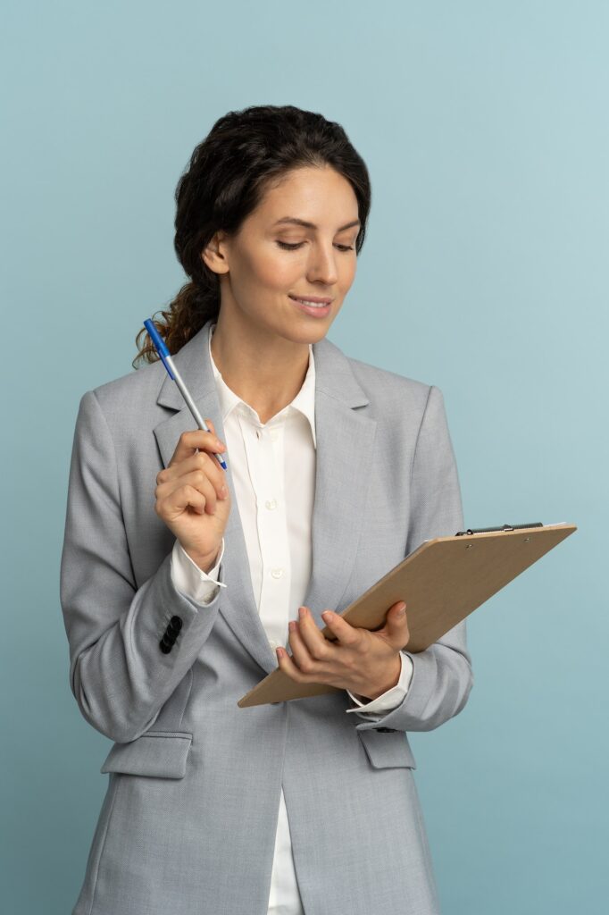 Friendly insurance agent woman wear grey blazer holding clipboard, pen looking at documents, studio.